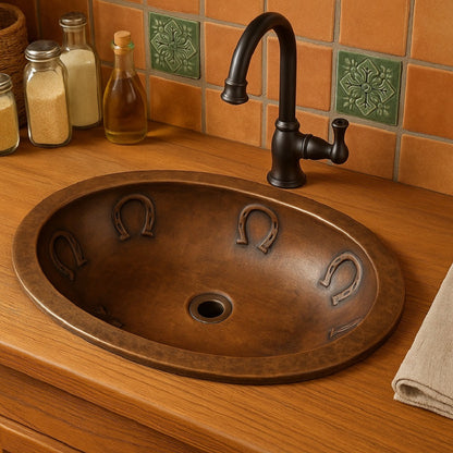 Custom oval copper drop in or undermount sink with horseshoe reliefs in a rustic bathroom with wooden counter and handmade ceramic tiles